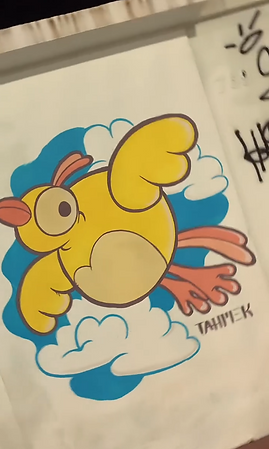 Grafite colorido de um pássaro amarelo de estilo cartunesco, com corpo arredondado, asas grandes e bico e pés laranjas. O pássaro aparece voando entre nuvens brancas contornadas em azul sobre fundo azul. No canto inferior direito está assinado 'TAHIMEK'. À direita há texto em grafite preto na parede.