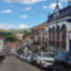 Rua histórica em Ouro Preto com casas coloniais e carros estacionados. Céu azul, colinas verdes ao fundo e decoração nas varandas dos sobrados.