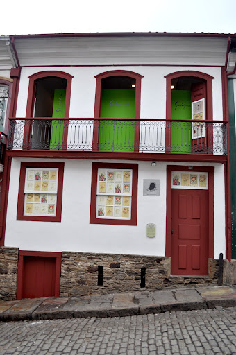 Edifício colonial de dois andares em rua de paralelepípedos inclinada. Fachada do museu Casa Guignard, branca com detalhes vermelhos nas janelas, portas e sacada. No andar superior, três aberturas em arco revelam paredes internas verdes e sacada com grade de ferro trabalhado. No térreo, porta vermelha à direita e janelas exibindo pôsteres coloridos. A base de pedra contrasta com a parte pintada.