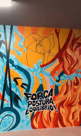 Mural colorido em parede com composição dinâmica de água e fogo. À esquerda, formas em tons de azul representam água; à direita, chamas vermelhas e laranjas simbolizam fogo. No centro, um desenho contínuo de rosto humano conecta os dois lados, sugerindo equilíbrio. Texto em letras pretas contornadas de branco diz 'FORÇA POSTURA EQUILÍBRIO!'.