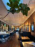 Bar with brick walls, marble counter, blue chairs, and a large plant. TVs on the wall, a few people sitting, and warm lighting overhead.