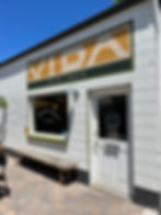 White bakery storefront with "Vida Baking Co" sign. "Gluten Free Bakery and Coffee" text on wall. Blue sky, rustic bench, "Open" sign on door.