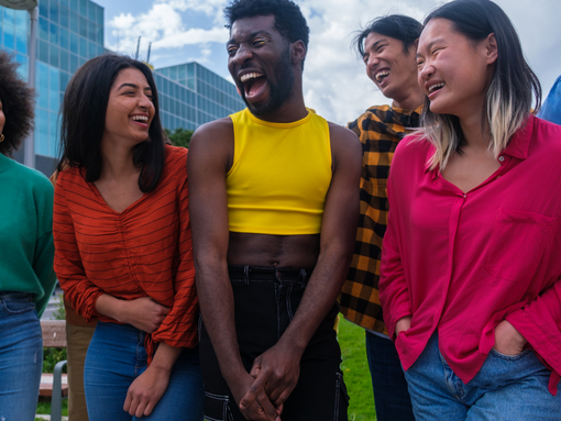 A diverse group of people laughing outdoors, wearing colorful outfits: green, red, yellow, plaid, and pink tops. Modern buildings in the background.