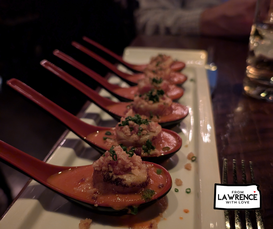 Red spoons with gourmet appetizers on a plate, garnished with herbs. Dimly lit restaurant table setting. Text: "From Lawrence with Love."