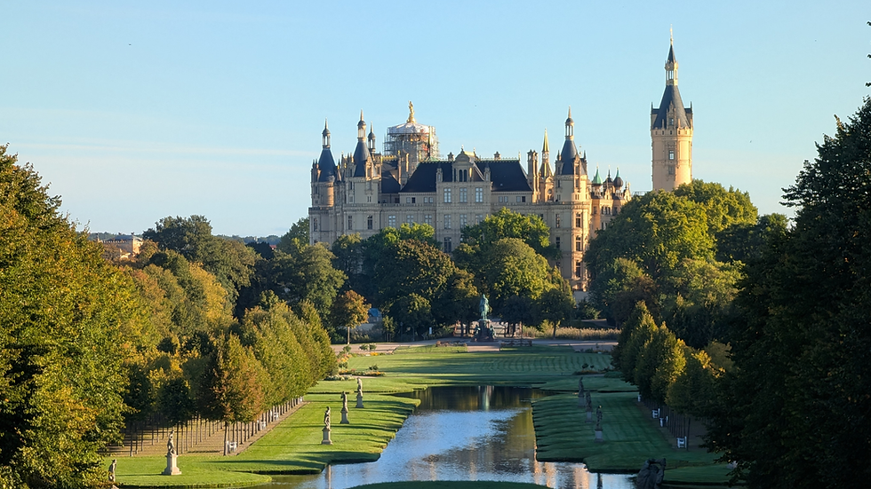 Sunlit fairytale castle with spires above formal gardens, canal, and statues, framed by lush trees under a clear blue sky.