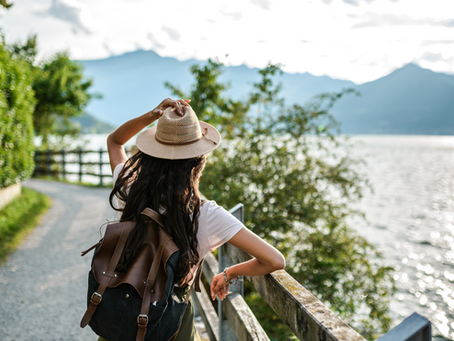 Woman in a hat and backpack stands on a lakeside path, gazing at the mountains. Sunlight reflects on the water, creating a serene mood.