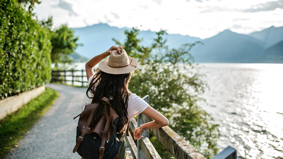 Woman in a hat and backpack stands on a lakeside path, gazing at the mountains. Sunlight reflects on the water, creating a serene mood.