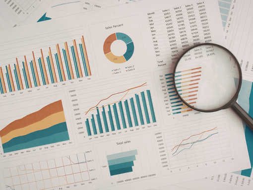 Charts and graphs with sales data on sheets in blue, orange, and teal. A magnifying glass is on top, emphasizing details.
