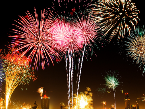 Colorful fireworks explode in the night sky over a city skyline, creating vibrant patterns of red, blue, and gold with an excited mood.