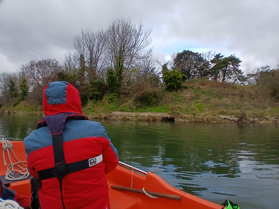 Image is of a person sitting in a small orange boat, taken from the perspective of another person sitting in the boat. Only the back of the person can  be seen, and they are wearing a red coat and flotation device. They are looking at a grassy bank with some trees on the side of the river.