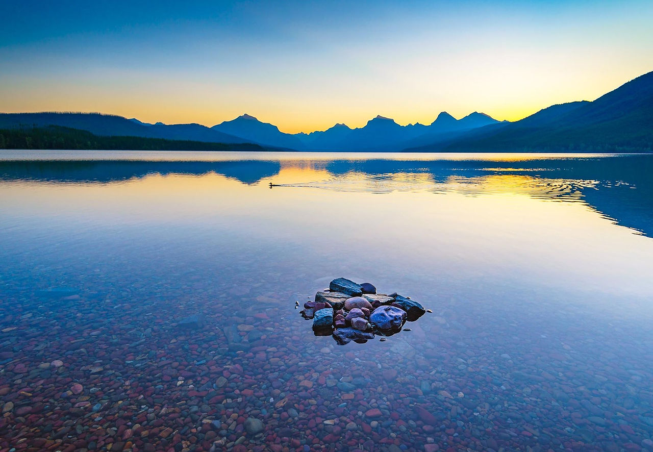 Still lake at sunrise with blue mountains and a small rock pile