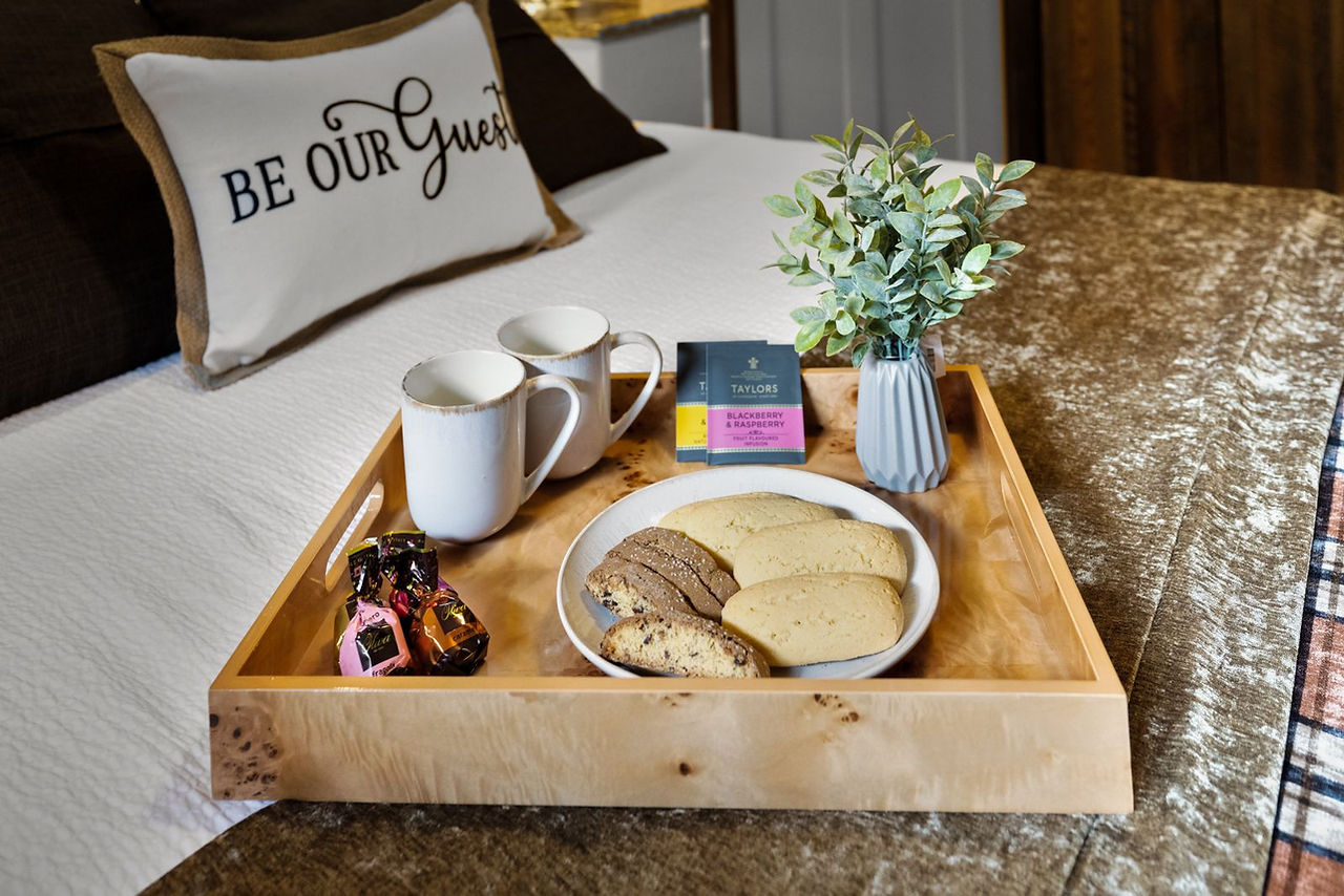 Somers House: a tray of mugs and cookies on the bed beside a “Be Our Guest” pillow
