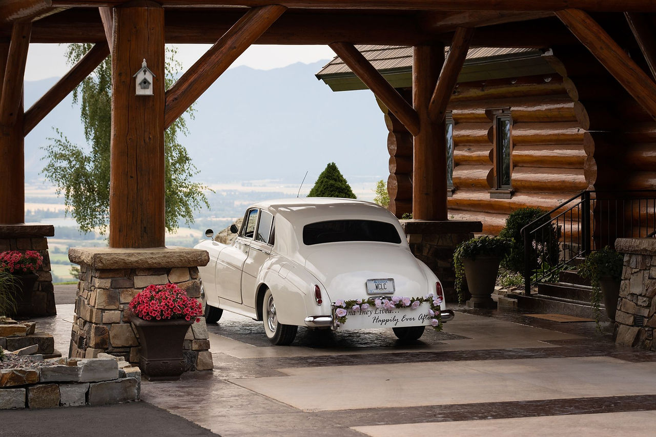 A white vintage car under the timber porte-cochere, lake beyond