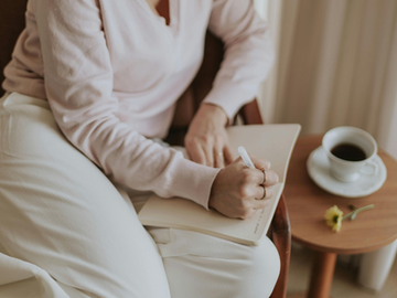 Woman sitting comfortably with a notebook, writing by hand beside a cup of coffee on a small table.