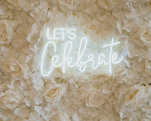 A chic 'Let's Celebrate' white neon sign for hire, mounted on a luxury ivory flower wall. The perfect backdrop for a baby shower, engagement party, or any special celebration.