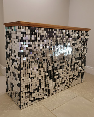 A bar unit covered in silver sequins featuring a glowing "Drinks" neon sign.
