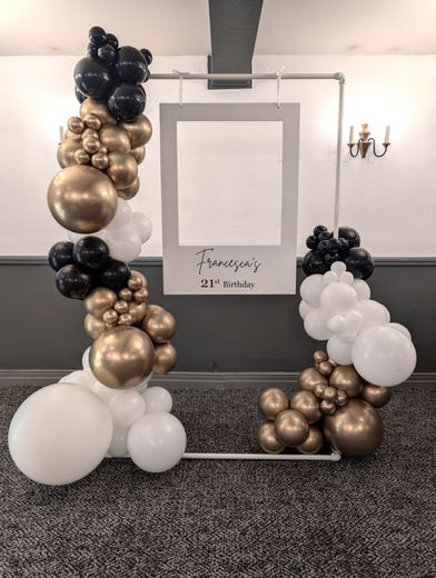 Modern photo booth backdrop with a white polaroid style frame and a black, white, and gold organic balloon garland.