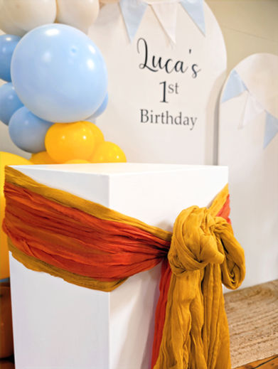 Modern and stylish first birthday party setup, including a white arched backdrop with personalised text, minimalist balloon decor, and a vibrant orange fabric drape over a white plinth.