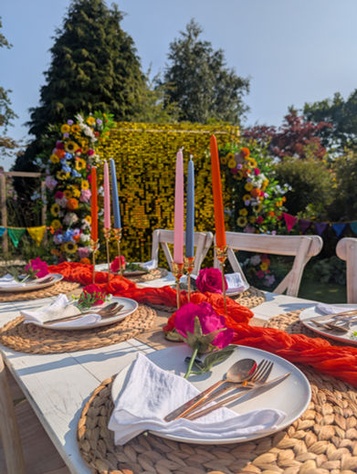Vibrant outdoor dining setup under a spectacular floral arch filled with bright red, pink, and orange flowers. The perfect statement decor hire for a festival wedding or garden party.