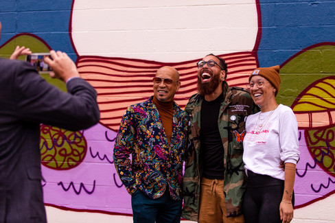 (left to right) Muralist for The 49507 Project, Erick Picardo, The Diatribe Executive Director Marcel Price, and Carnevale Director of Social Impact Nika Price pose for a smartphone picture in front of Tony WHLGN's mural on The Old Goat.