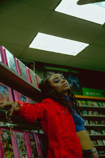 Model with hair in long ponytails leans against a wall of VHS tapes. There is a vintage feel to the image.