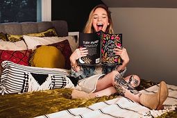 Business owner poses with her product, an illustrated coloring book. She is tattooed and excited, and sits on a bohemian-style bed.