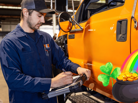Mechanic in a blue uniform writes on a clipboard beside an orange truck in a garage. A pot of gold, rainbow, and clovers overlay the image.