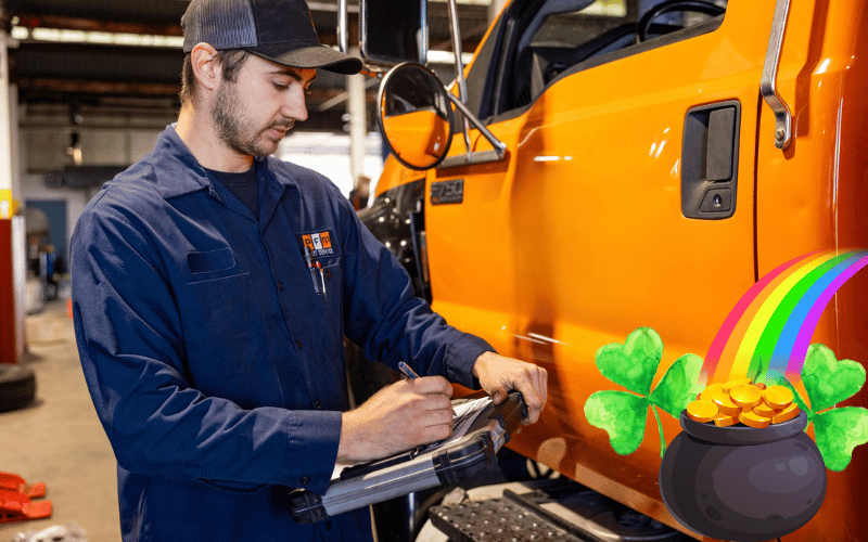 Mechanic in a blue uniform writes on a clipboard beside an orange truck in a garage. A pot of gold, rainbow, and clovers overlay the image.