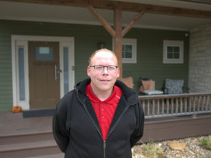 A man with short red hair wears glasses, a red shirt, and a black hoodie. He stands in front of a house with a large porch.