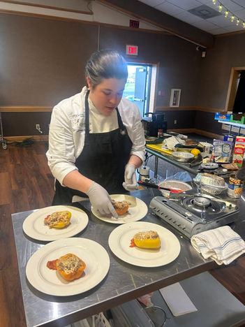 A person wearing a chef's coat and apron cooks over a portable stove on a stainless steel prep table, with ingredients and kitchenware visible.