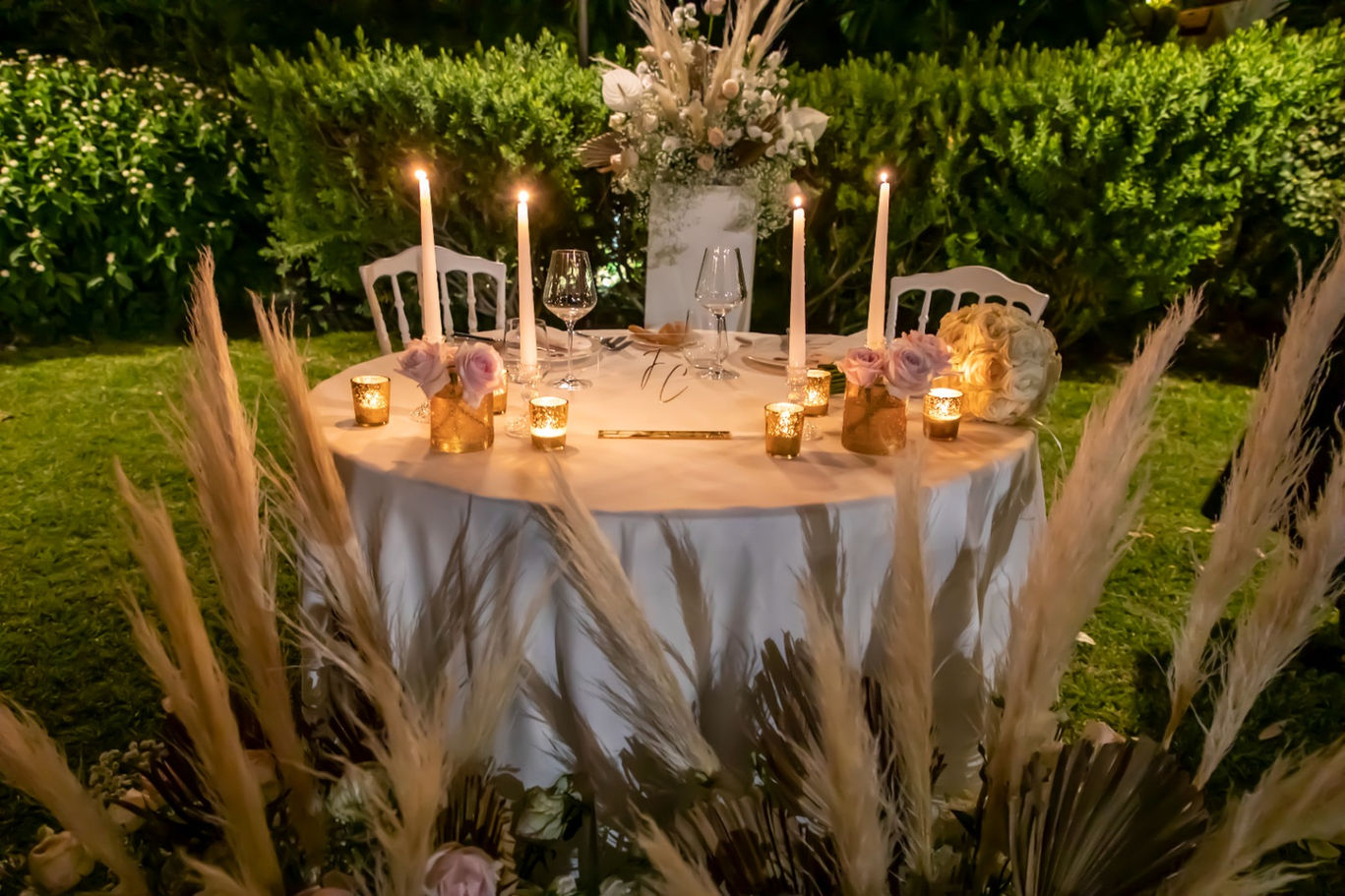 Sofisticato allestimento bianco e oro del tavolo degli sposi (ristorante) in giardino. Con centrotavola di fiori, candele e pampas e alla base addobbi floreali di rose bianche, rosa, gialle, e fiorellini di campo.