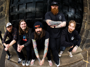 Five men with tattoos sit on steps, casually posed, in front of a large wooden door. They wear casual clothes and sneakers. Mood is relaxed.
