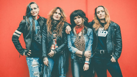 Four women in leather jackets and band tees stand against a bright red wall, exuding a confident and rebellious vibe.