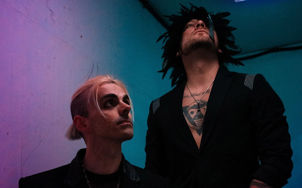 Two men with dark clothing, one with blond hair, the other with spiky black hair, look up in a dimly lit room with colorful lighting.