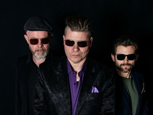 Three men wear sunglasses against a dark background. They display a cool, confident vibe. One wears a purple shirt and patterned jacket.