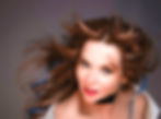Woman with flowing brown hair, hoop earrings, and red lipstick gazes at camera. Background is softly blurred, giving a dramatic feel.