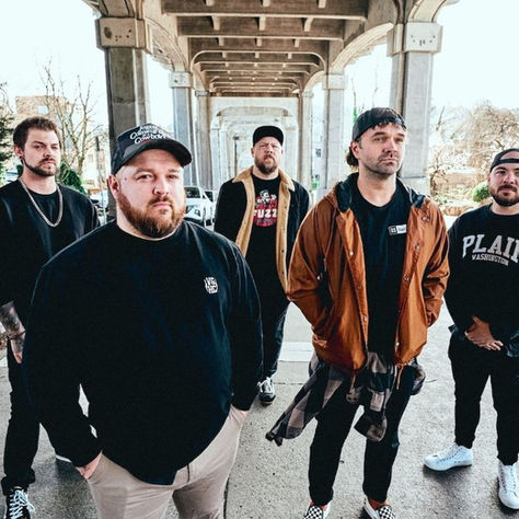 Five men pose under a concrete overpass, one in an orange jacket and one shirt reading PLAIN WASHINGTON, with a cool urban mood.