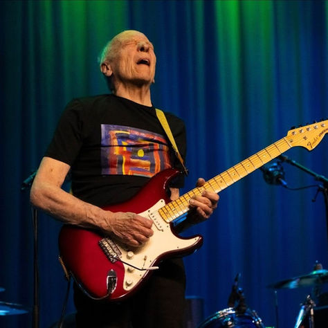 Elderly man passionately plays a red electric guitar on stage with a vivid blue and green curtain backdrop, conveying a lively mood.