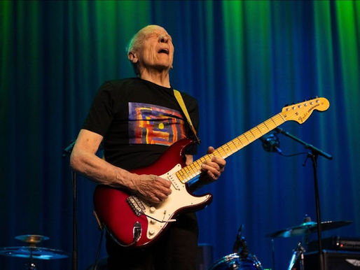 Elderly man passionately plays a red electric guitar on stage with a vivid blue and green curtain backdrop, conveying a lively mood.
