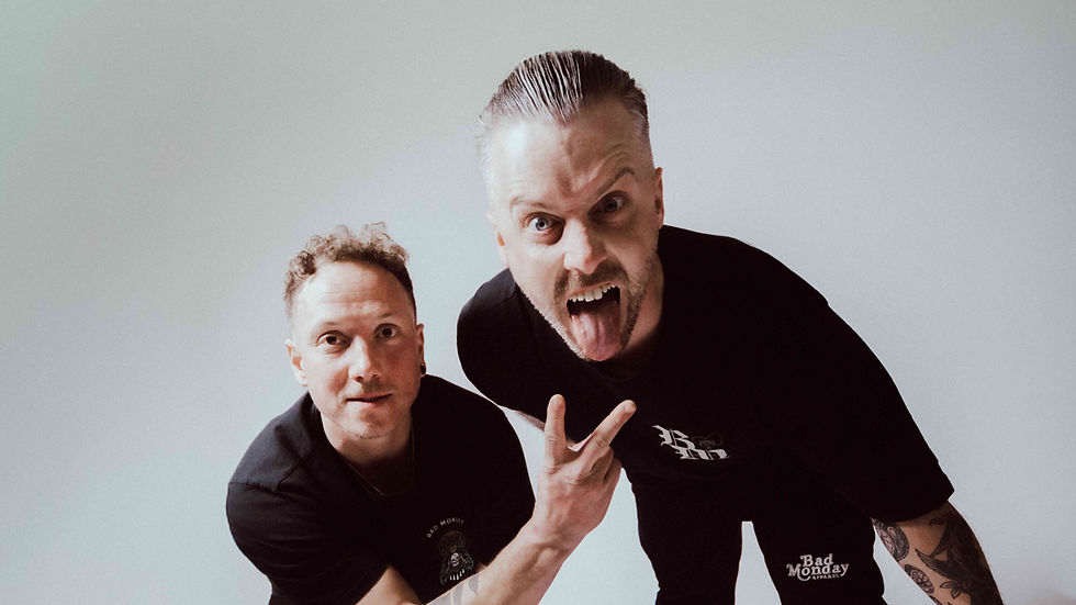 Two men in black shirts pose against a plain white background; one sticks out his tongue while the other flashes a peace sign.