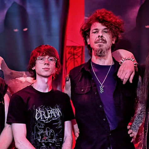 Four men pose under red-purple stage lights; one wears a Brujeria shirt, with dark band posters behind them.
