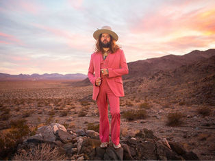 Man in pink suit and hat stands on rocky desert terrain at sunset, with mountains in background. Sky glows with orange and pink hues.