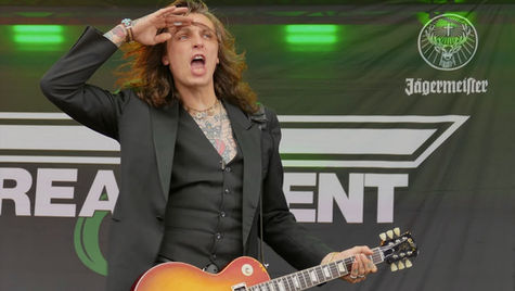 Guitarist in black suit passionately performing on stage, saluting with right hand. Background shows Jägermeister branding, vibrant mood.