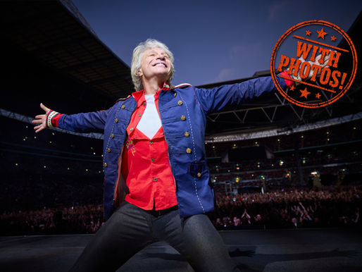 Smiling performer in blue and red jacket spreads arms on a stadium stage; orange stamp reads WITH PHOTOS!