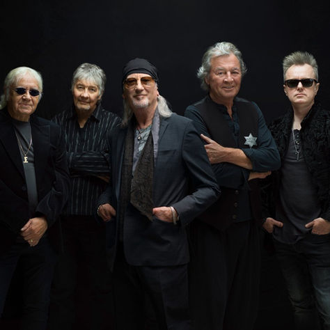 Five older men in black outfits and sunglasses pose against a black studio background, looking serious and stylish.