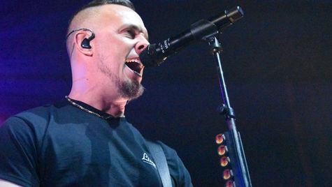 A man passionately sings into a microphone on stage, wearing a black shirt. The background is dimly lit, creating a dramatic atmosphere.
