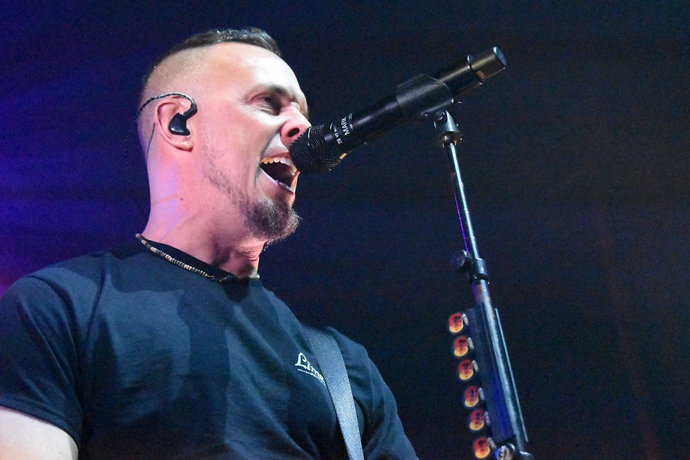 A man passionately sings into a microphone on stage, wearing a black shirt. The background is dimly lit, creating a dramatic atmosphere.