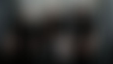 Four people in black outfits stand confidently against a wooden backdrop. Tattoos and accessories add to their edgy style. Moods are serious.