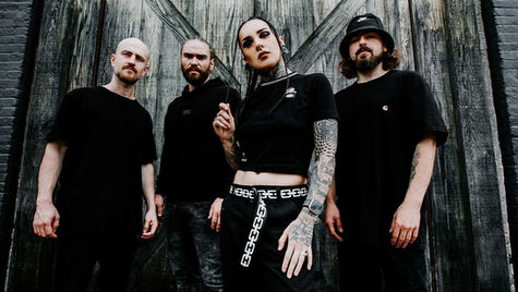 Four people in black outfits stand confidently against a wooden backdrop. Tattoos and accessories add to their edgy style. Moods are serious.