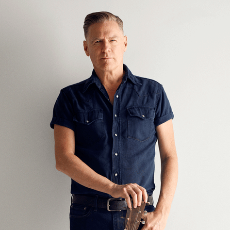 Man in a navy shirt stands against a white wall, resting a hand on a guitar with a serious expression.